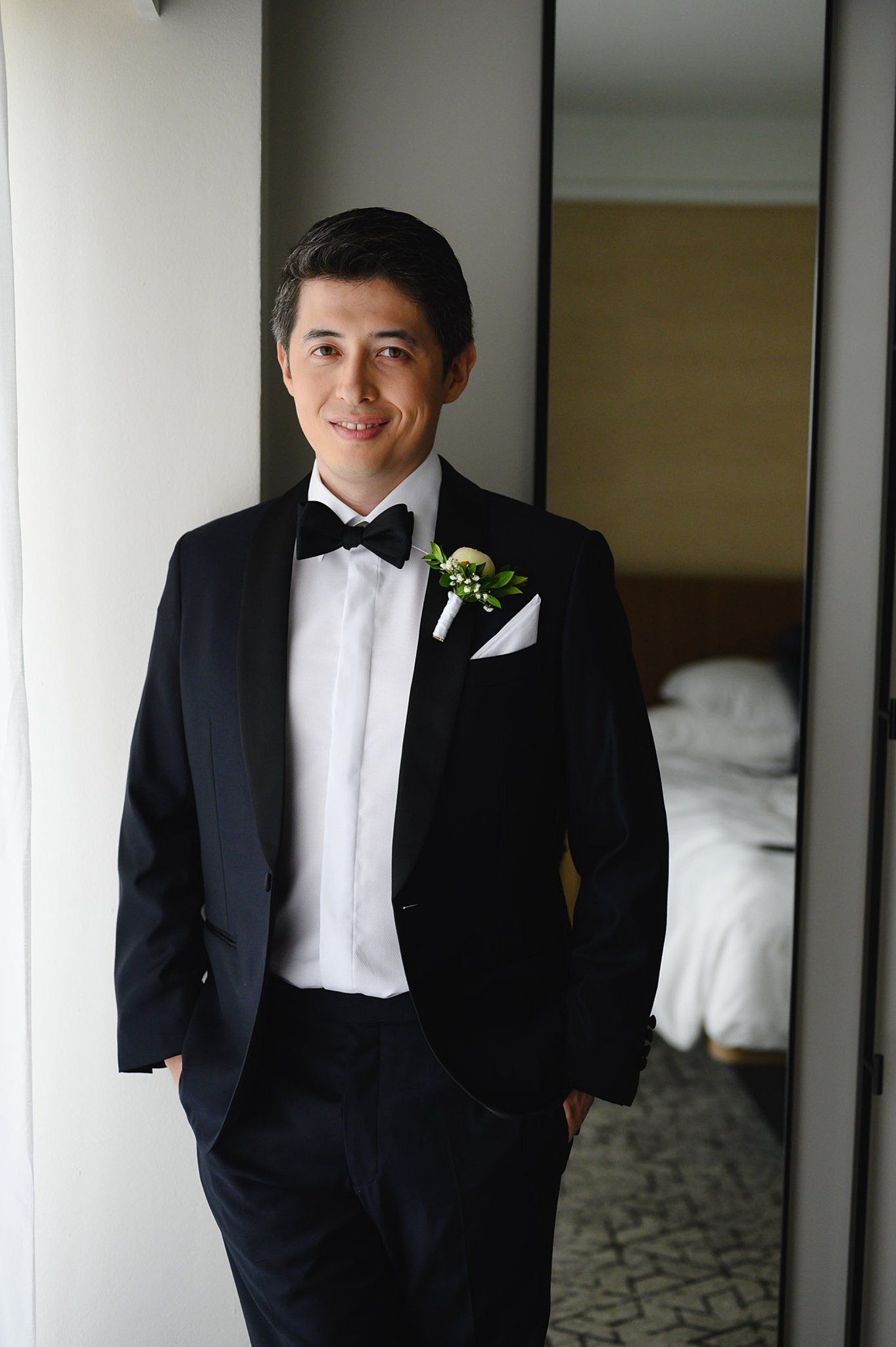 Groom in black tuxedo with bow tie and boutonniere smiling in hotel room before wedding ceremony.