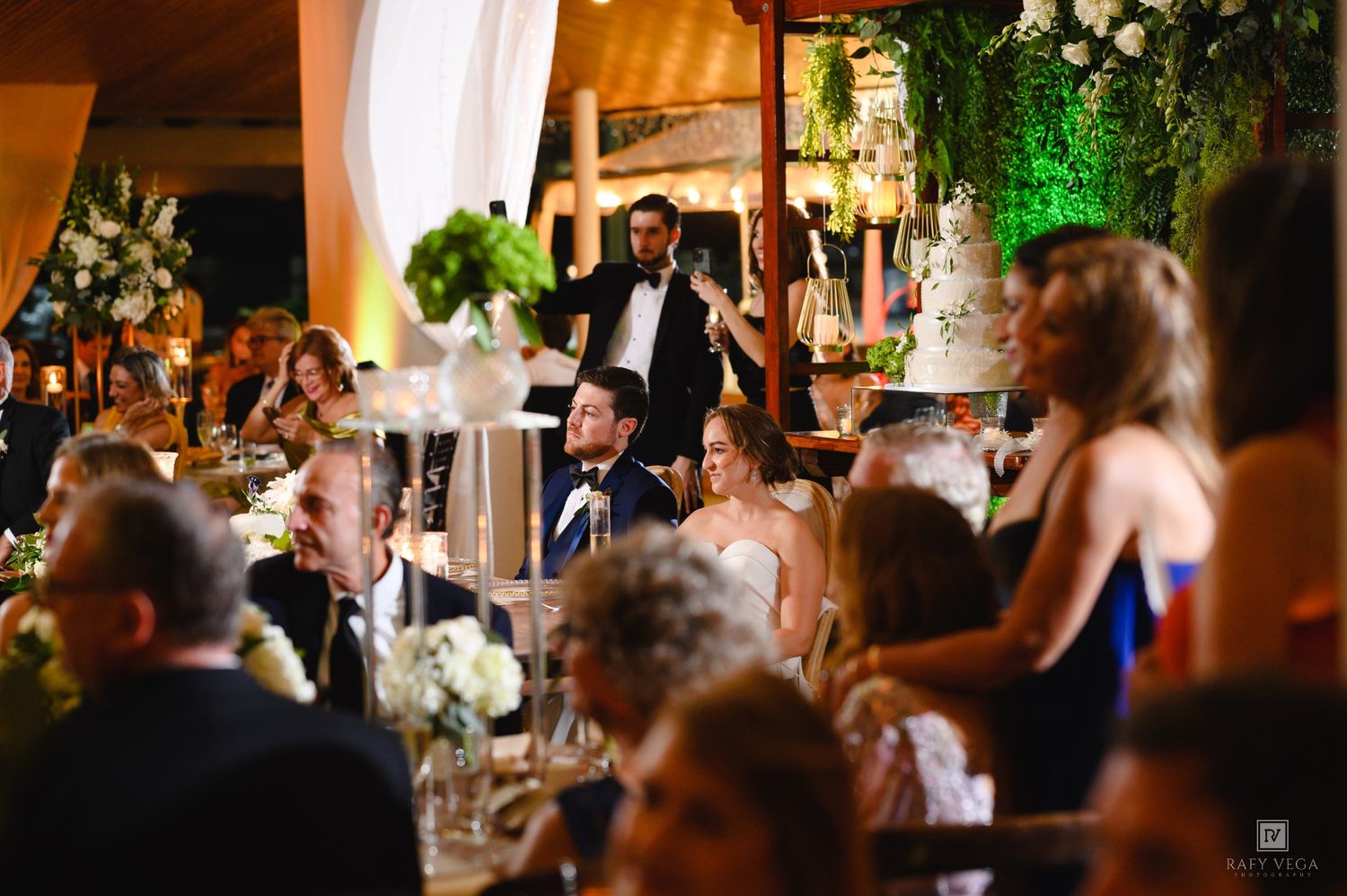 A man gives a toast speech at an elegant wedding reception with floral arrangements and a tiered cake in the background.
