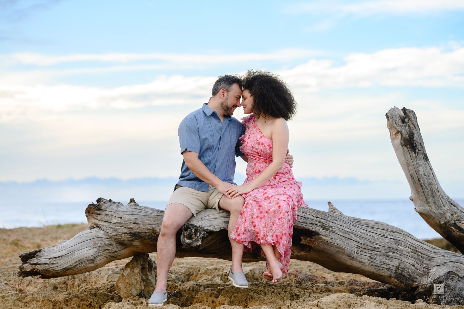 Romantic couple sharing a tender moment sitting on driftwood at a scenic beach with ocean views.