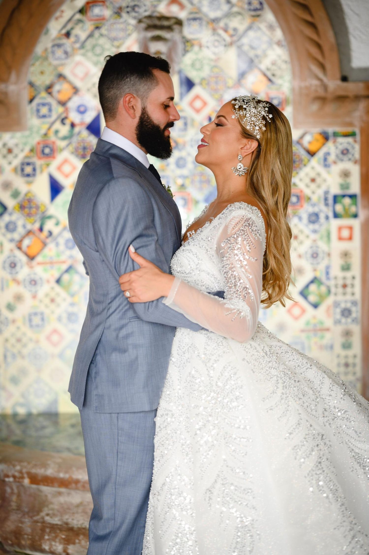 A bride and groom share a romantic moment in front of a colorful mosaic tile wall, dressed in wedding attire.