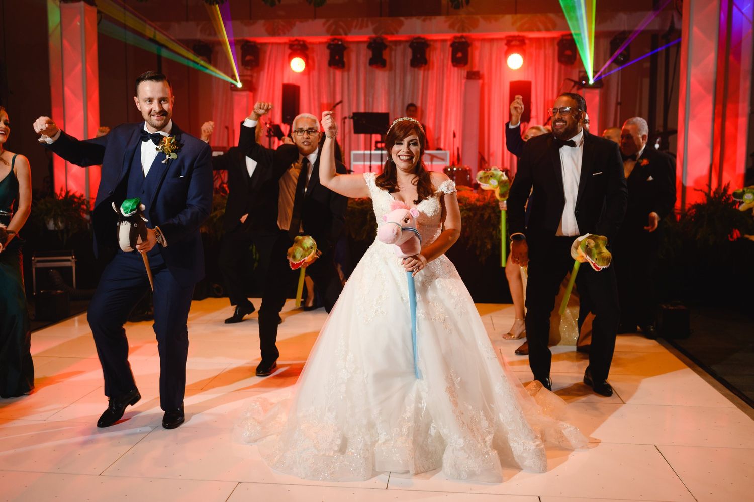 Bride in white ball gown celebrates with groomsmen in navy suits on a wedding reception dance floor.