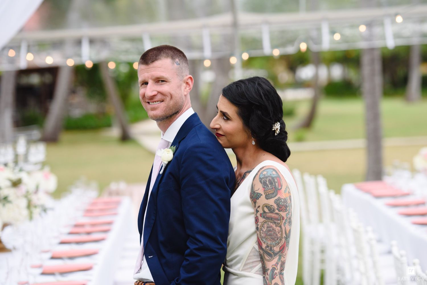 Bride with floral tattoo sleeve and groom in navy suit smile at outdoor wedding reception with string lights.
