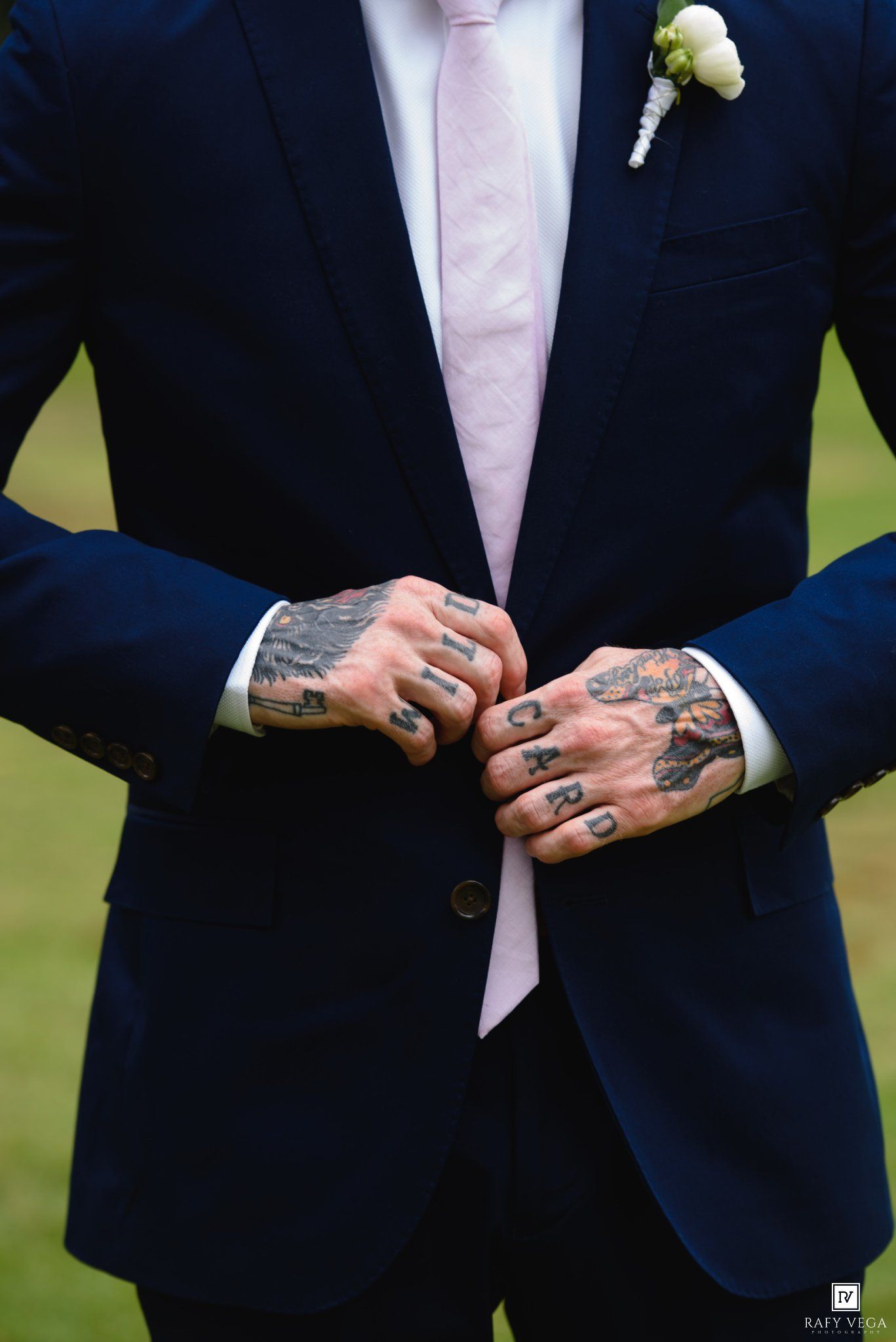 Groom in navy suit with pink tie and boutonniere, showing heavily tattooed hands clasped together.