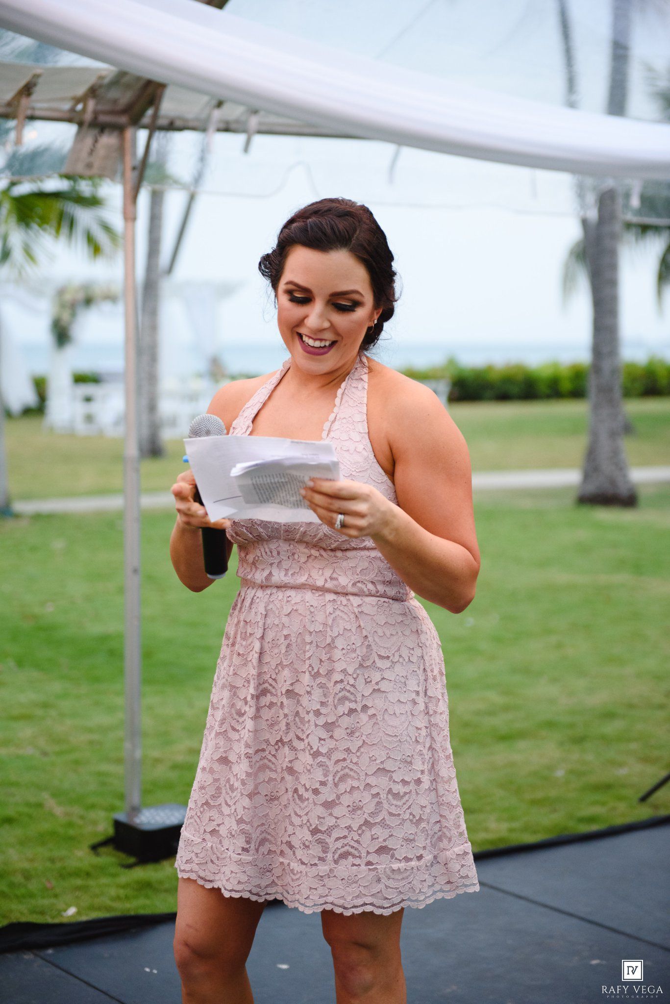 A bridesmaid in a pink lace dress reads a heartfelt speech at an outdoor wedding ceremony by the ocean.