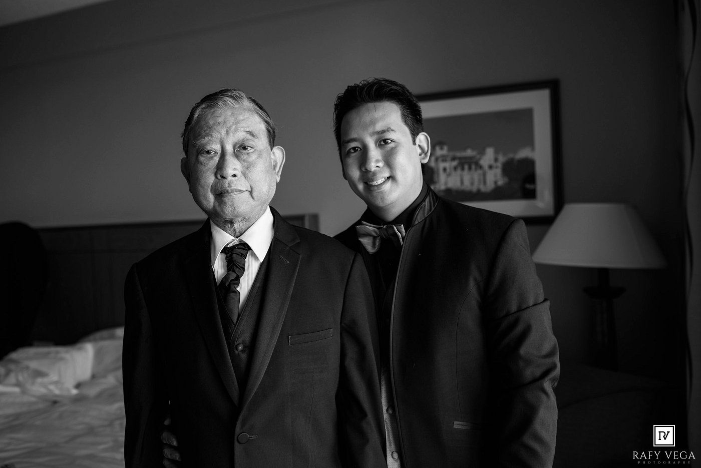 Black and white photo of two men in suits posing together in a hotel room at a wedding.