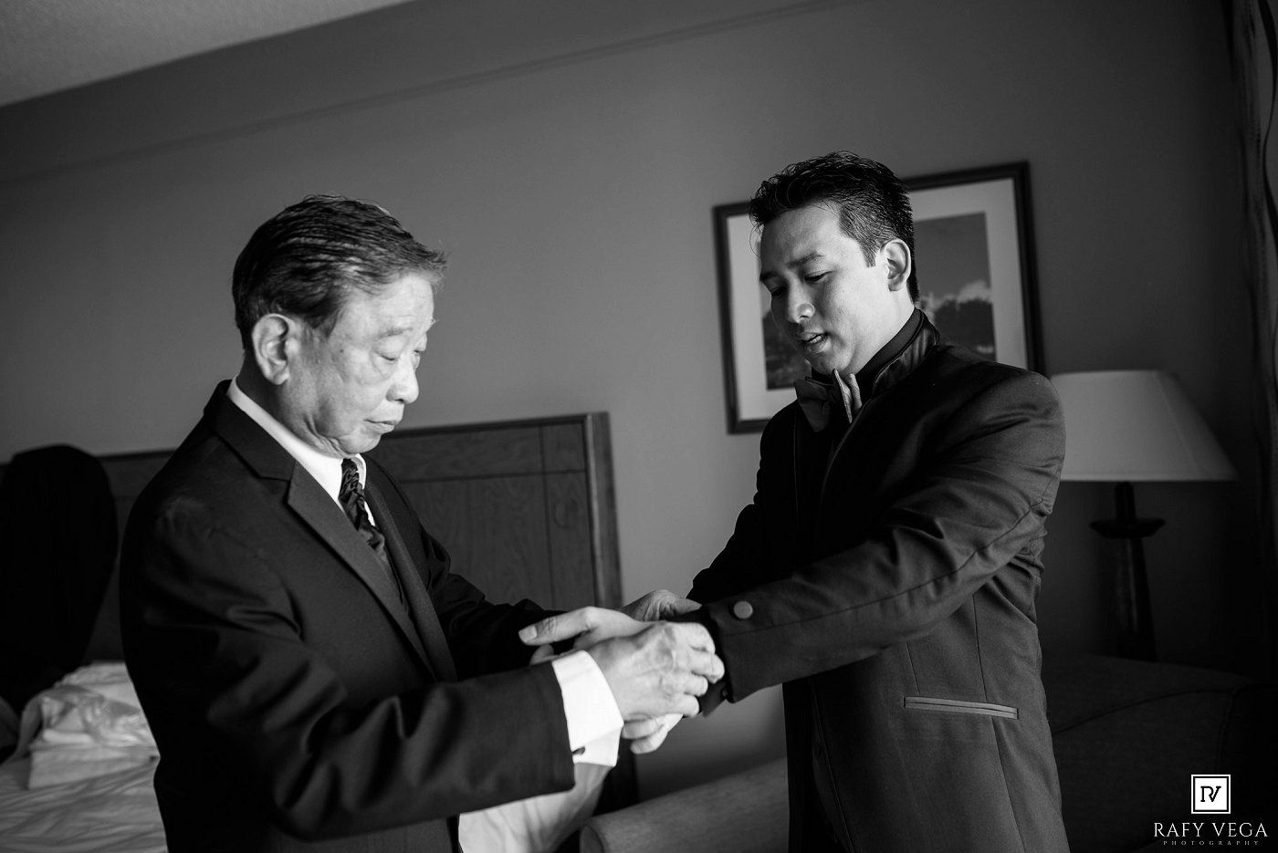 Black and white photo of two men in suits, one helping the other with his cufflinks in a hotel room.
