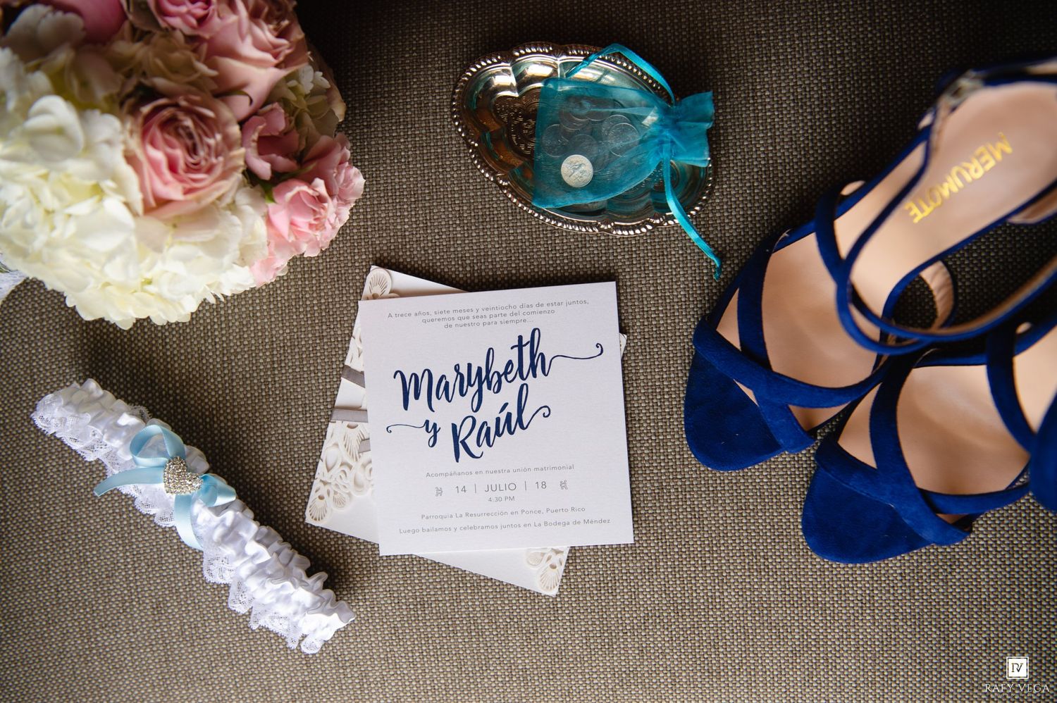 Blue bridal heels, wedding invitation for Marybeth and Raul, lace garter, and pink flowers arranged on a neutral surface.