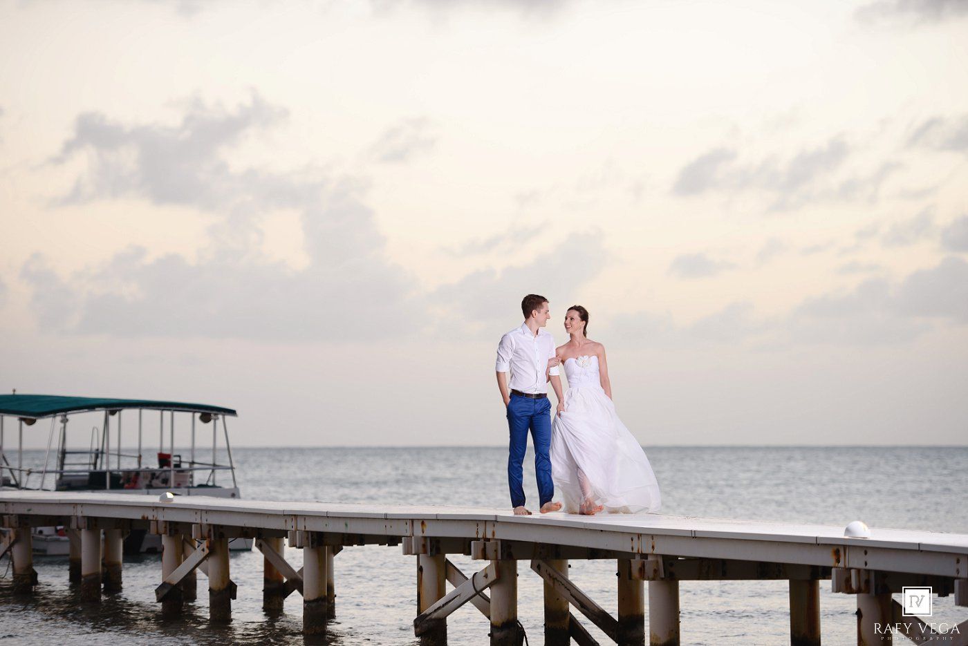 Bride and groom stand on a wooden pier over calm ocean waters at sunset in a romantic beach wedding photo.