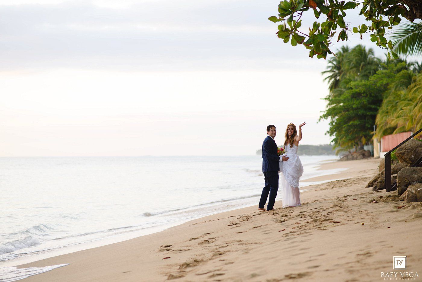 Couple in wedding attire posing on a tropical beach at sunset with lush palm trees and calm ocean waves.