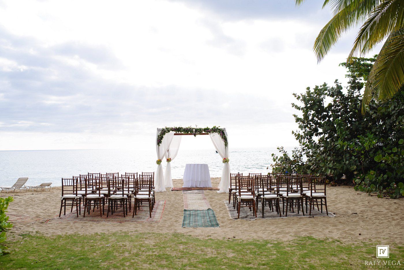 Beach wedding ceremony setup with wooden chairs, floral arch, and ocean backdrop on sandy shore.