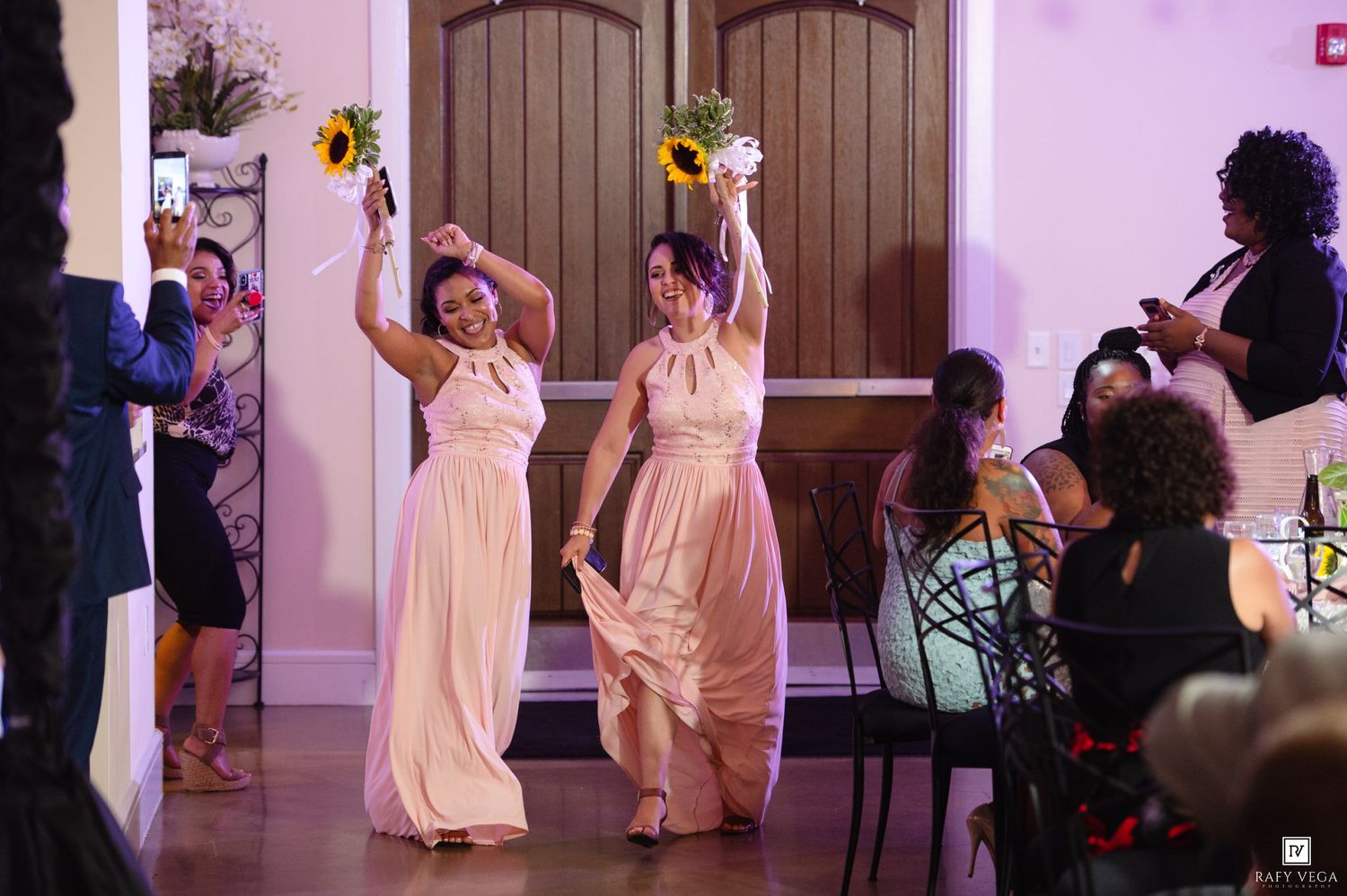 Two bridesmaids in blush pink dresses raise sunflower bouquets at wedding reception.