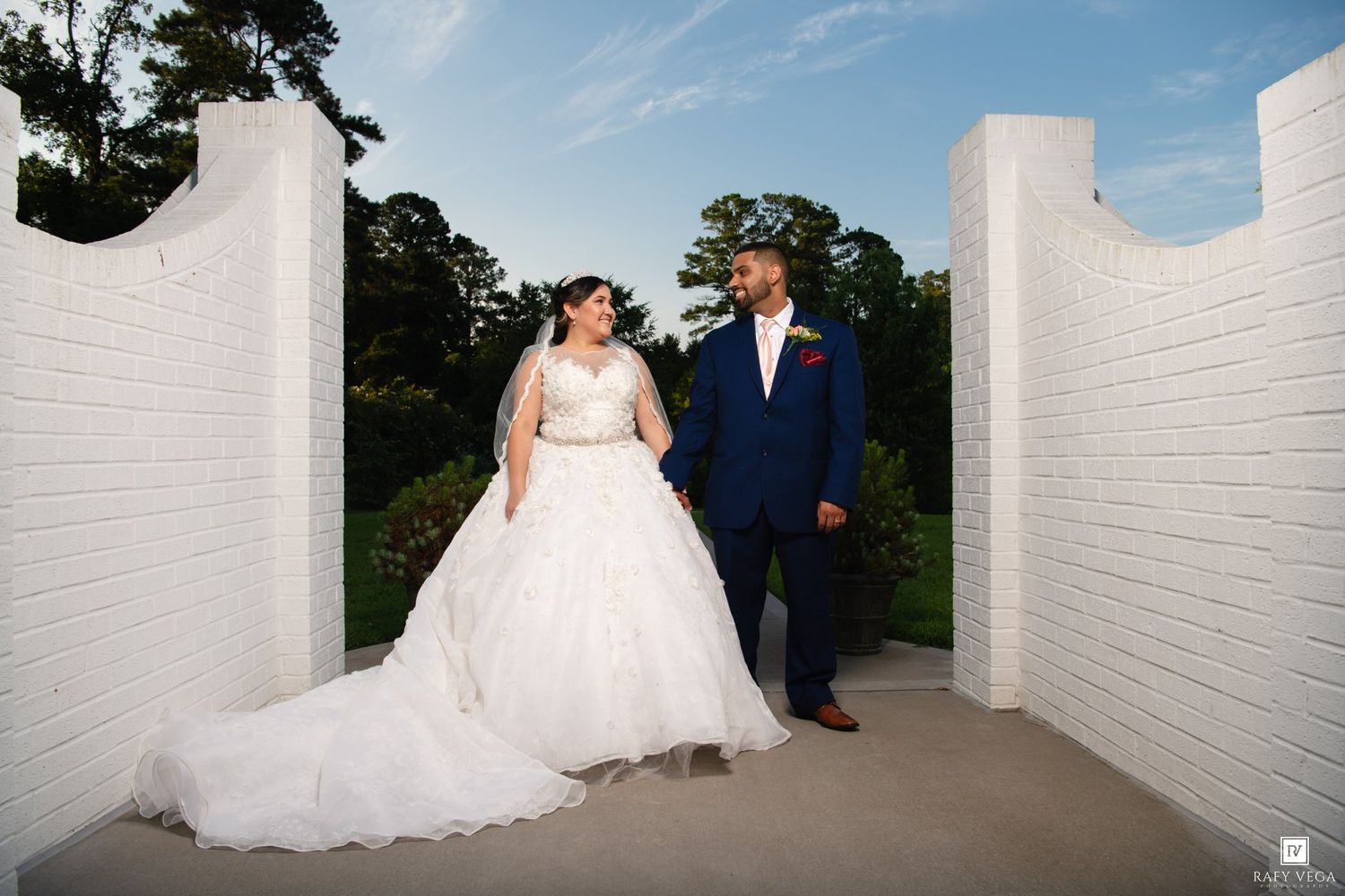 Bride in white ball gown and groom in navy suit stand between white brick pillars at outdoor wedding venue.