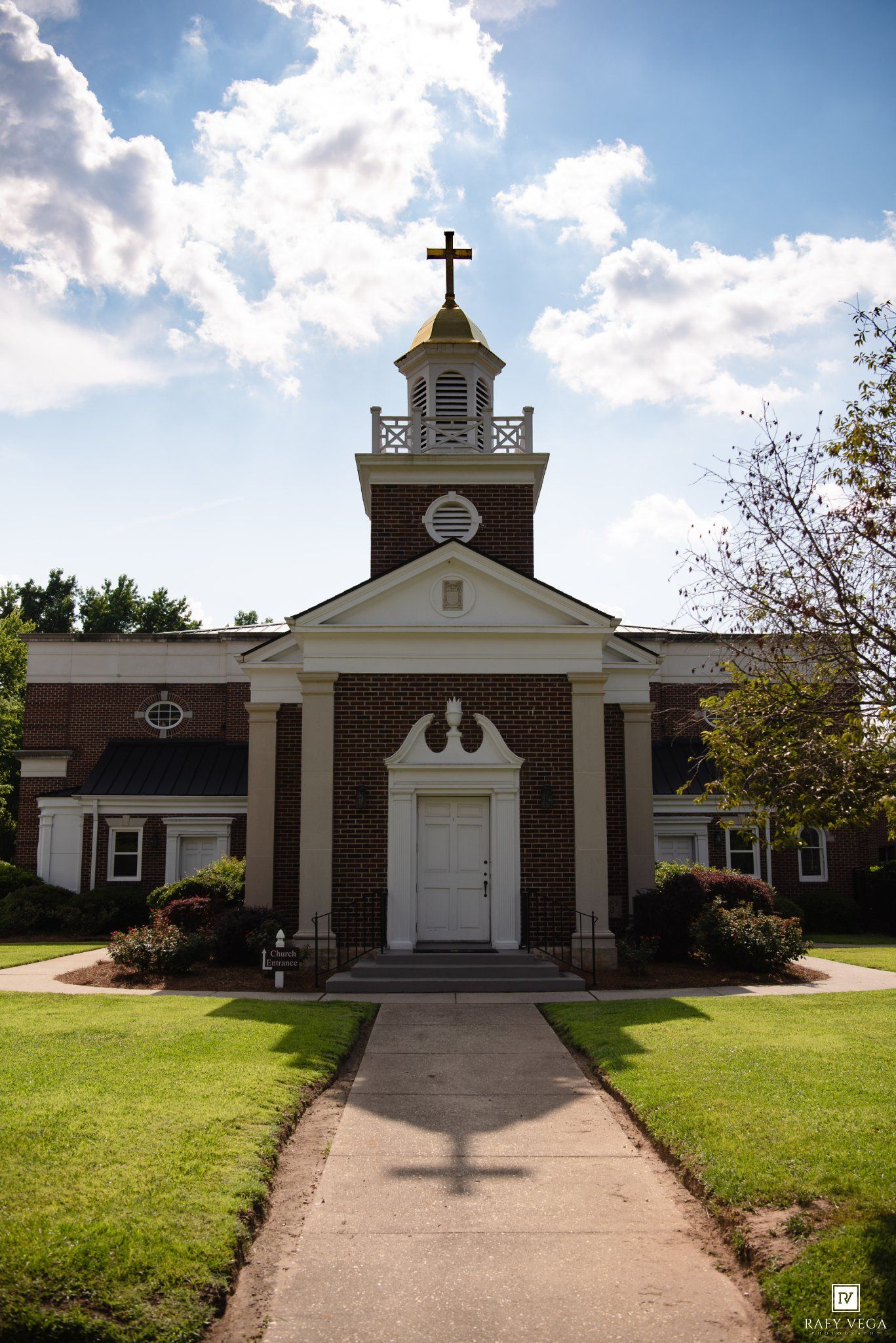 Brick church with white columns, steeple cross, and golden dome under blue sky with clouds and tree-lined pathway.