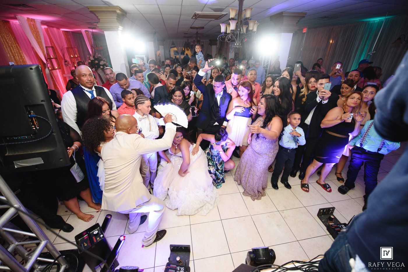 Bride and groom celebrate on a crowded wedding reception dance floor surrounded by guests and colorful lights.