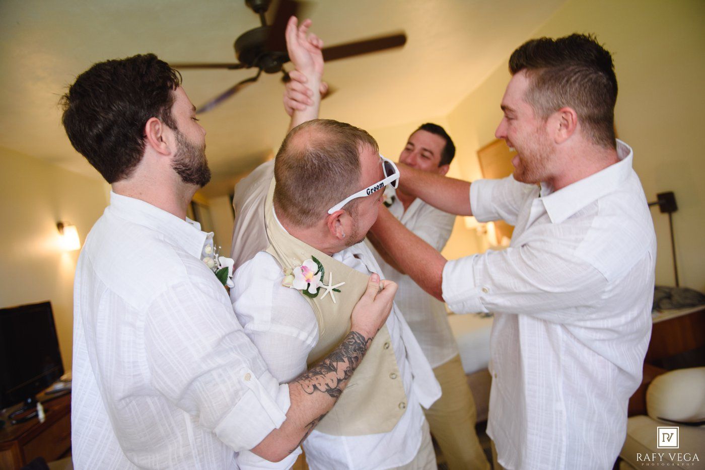Groomsmen helping groom get dressed in white shirts and vest before wedding ceremony indoors.