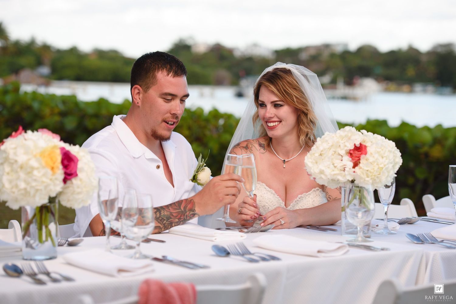 Bride and groom toasting with champagne at outdoor waterfront wedding reception table with floral centerpieces.
