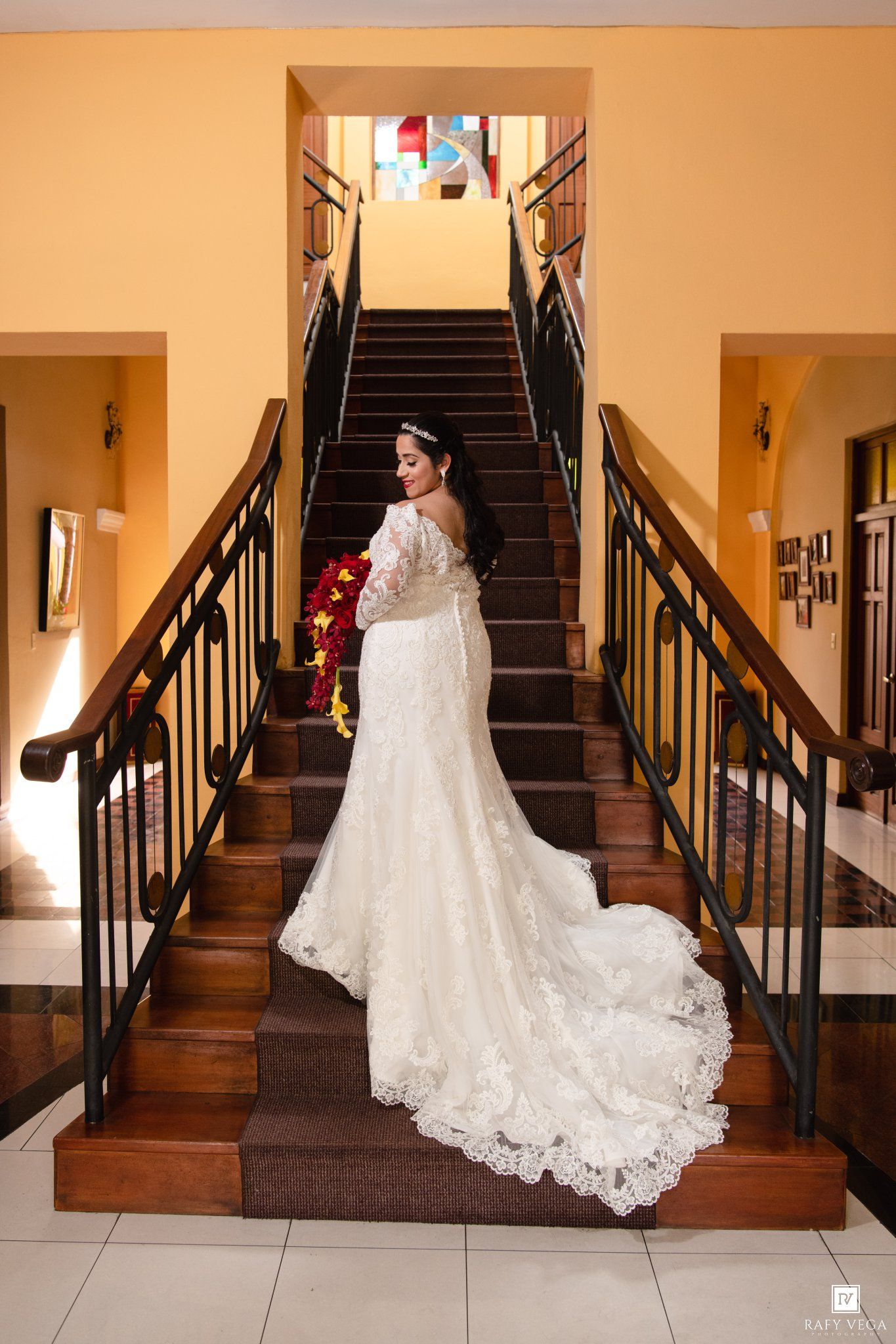 Bride in elegant lace mermaid wedding gown with long train stands on wooden staircase holding red bouquet.