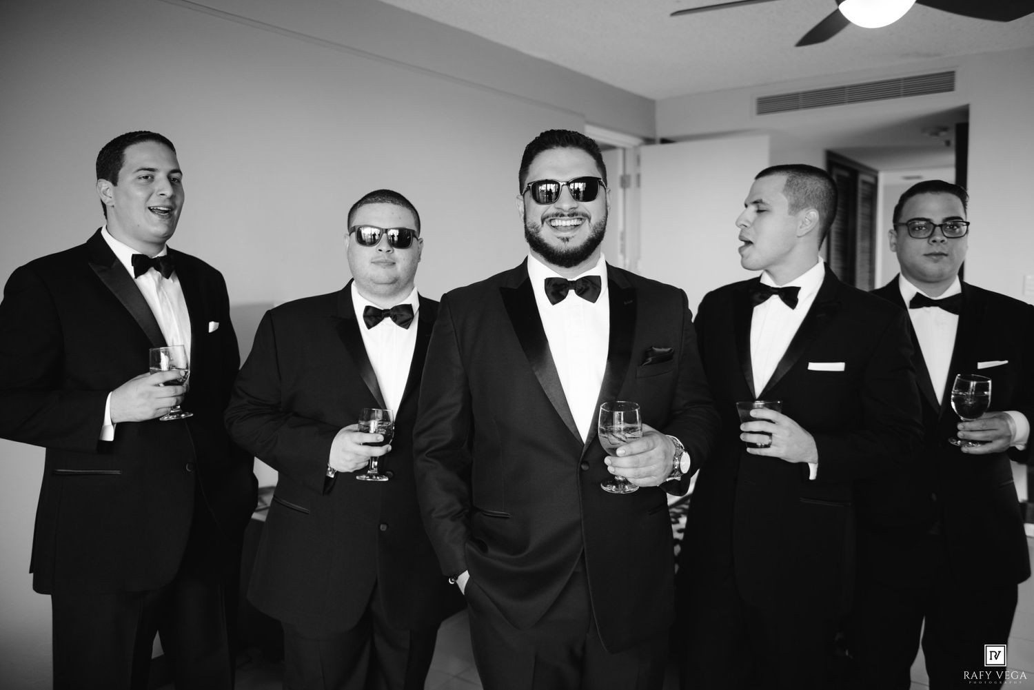 Groom and groomsmen in tuxedos holding drinks and smiling in black and white wedding photo.