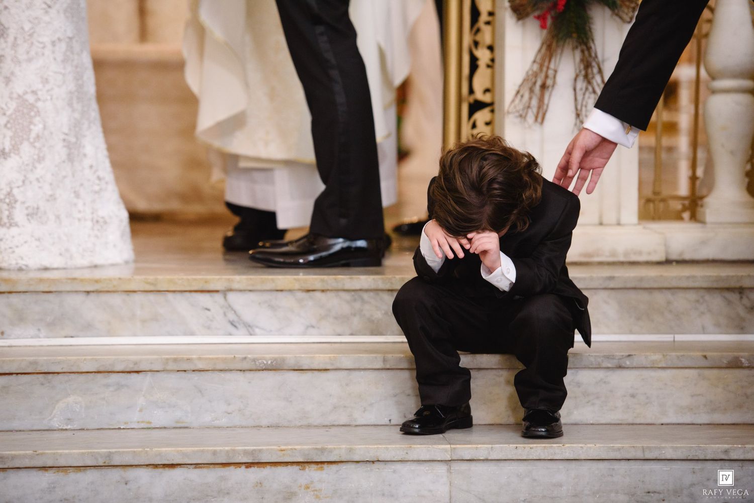 Young boy in black suit sitting on church steps with head down during a wedding ceremony.