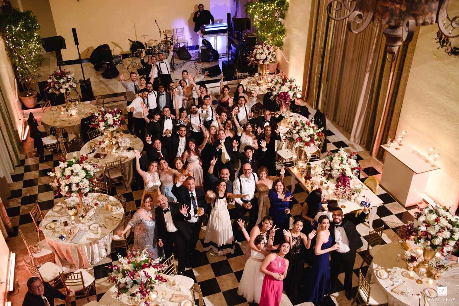 Aerial view of wedding reception guests posing together on checkered dance floor surrounded by floral centerpieces.