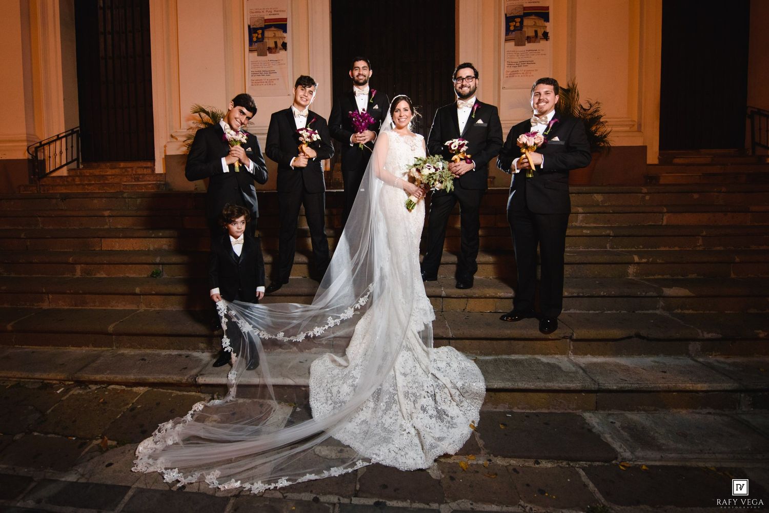 Wedding party poses on church steps, bride in lace gown with long veil, groom and groomsmen in black suits.
