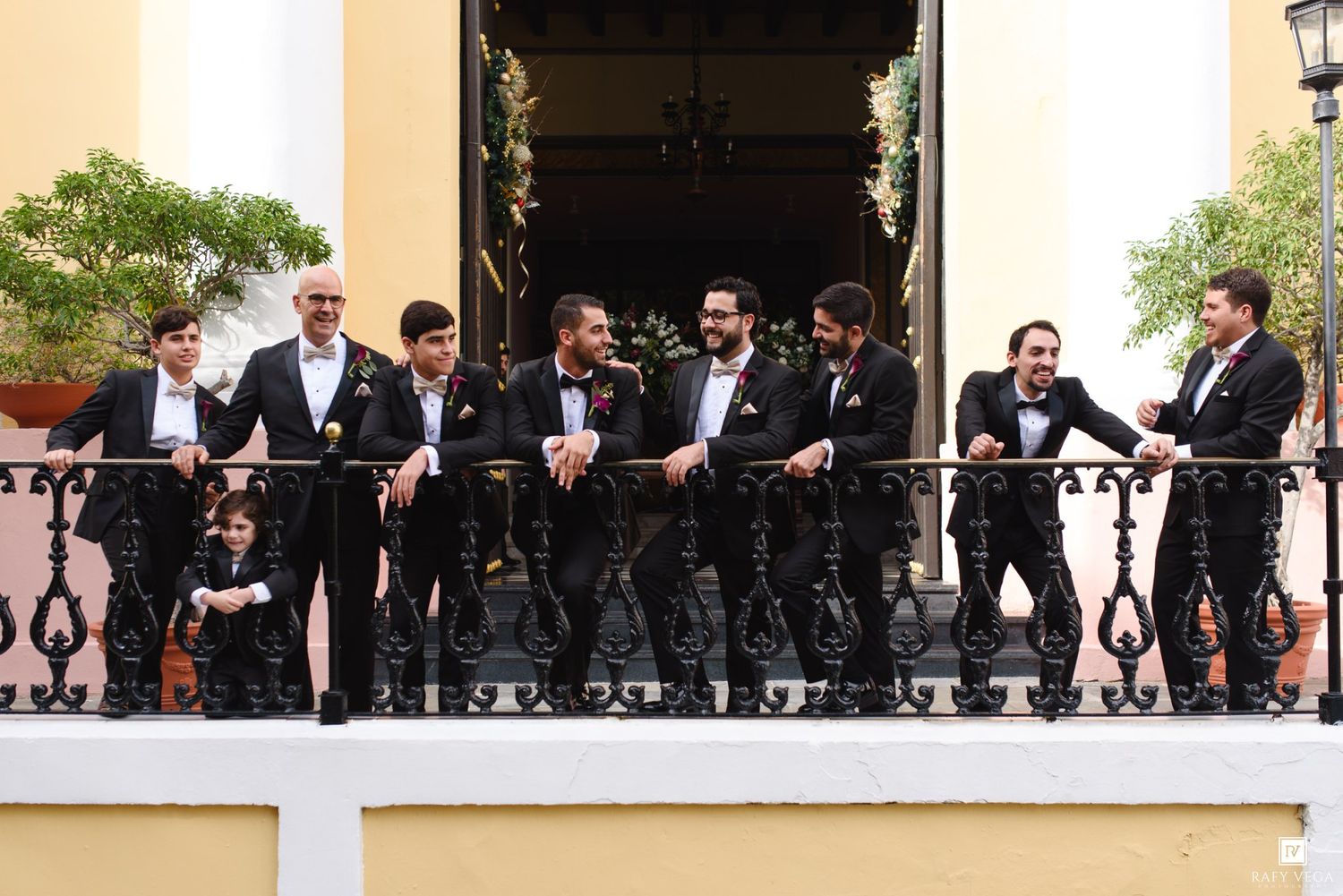 Groomsmen in black tuxedos with bow ties posing on an ornate balcony railing at a wedding venue.