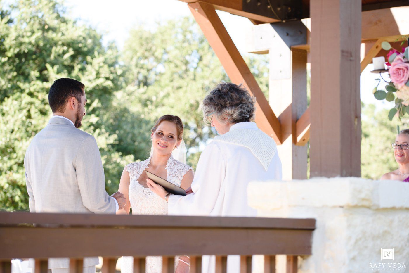Bride and groom exchange vows with officiant at outdoor wedding ceremony under wooden pavilion.