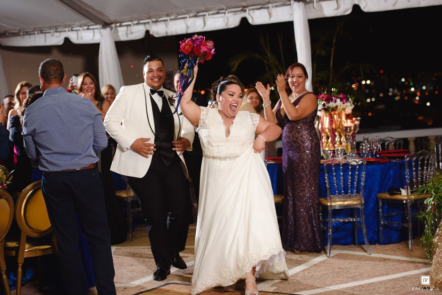 Bride and groom make joyful grand entrance at elegant outdoor wedding reception, bride holding colorful bouquet aloft.