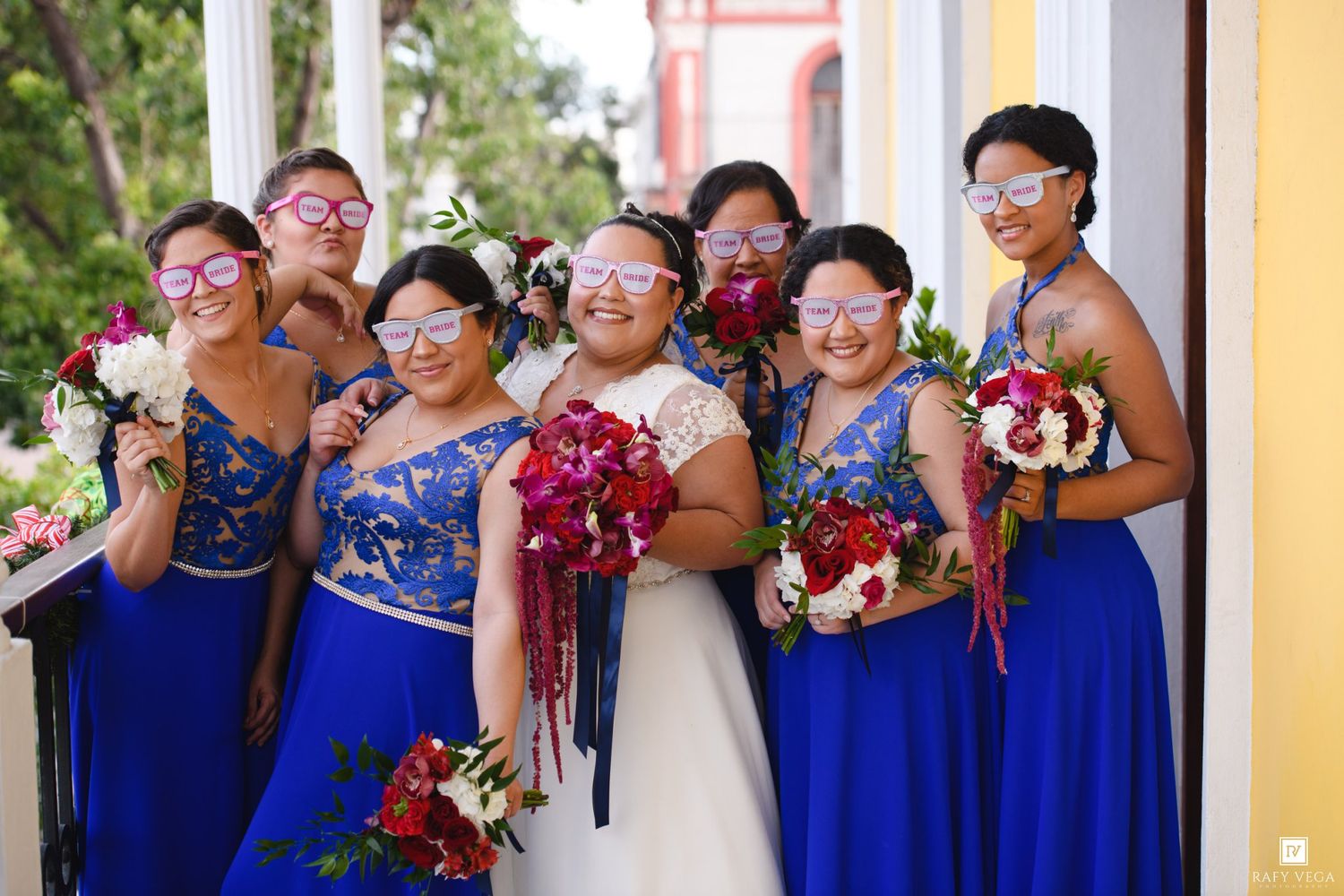 Bride and bridesmaids in royal blue dresses wearing fun sunglasses holding bouquets at an outdoor wedding celebration.