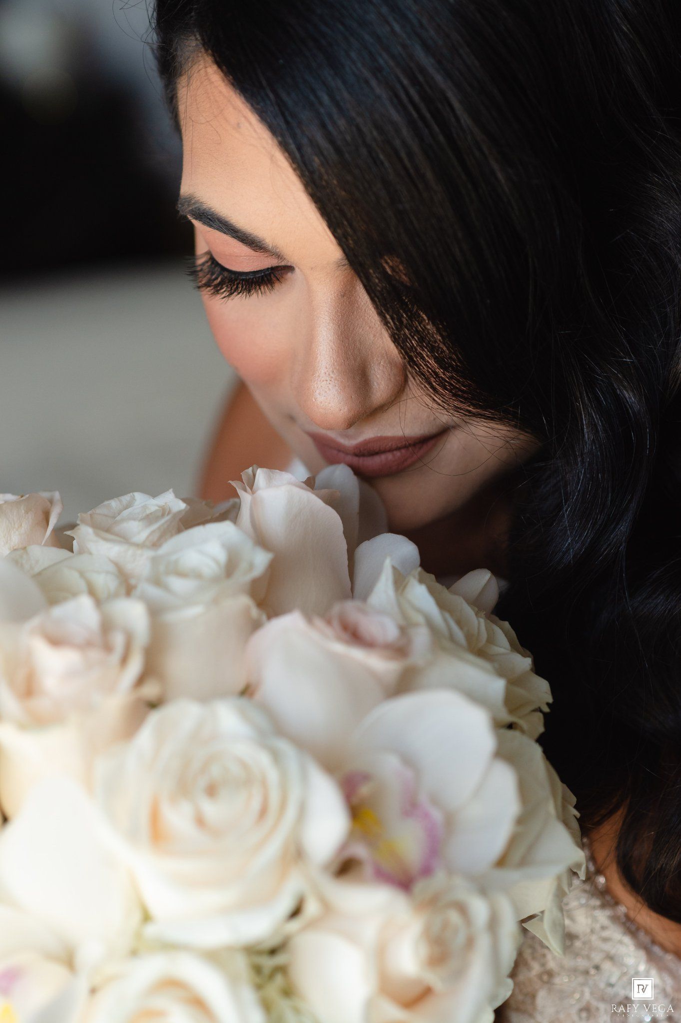Bride smelling a lush bouquet of white and blush roses with elegant makeup and dark wavy hair.
