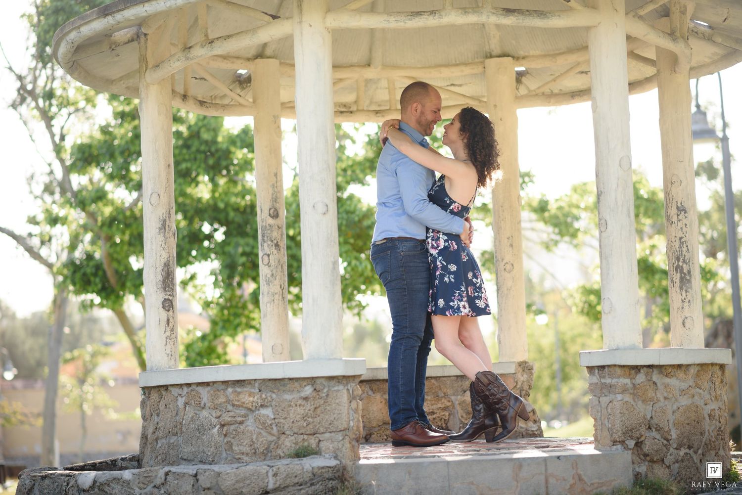 Couple embracing under a white gazebo with columns, surrounded by lush green trees, in a romantic outdoor setting.