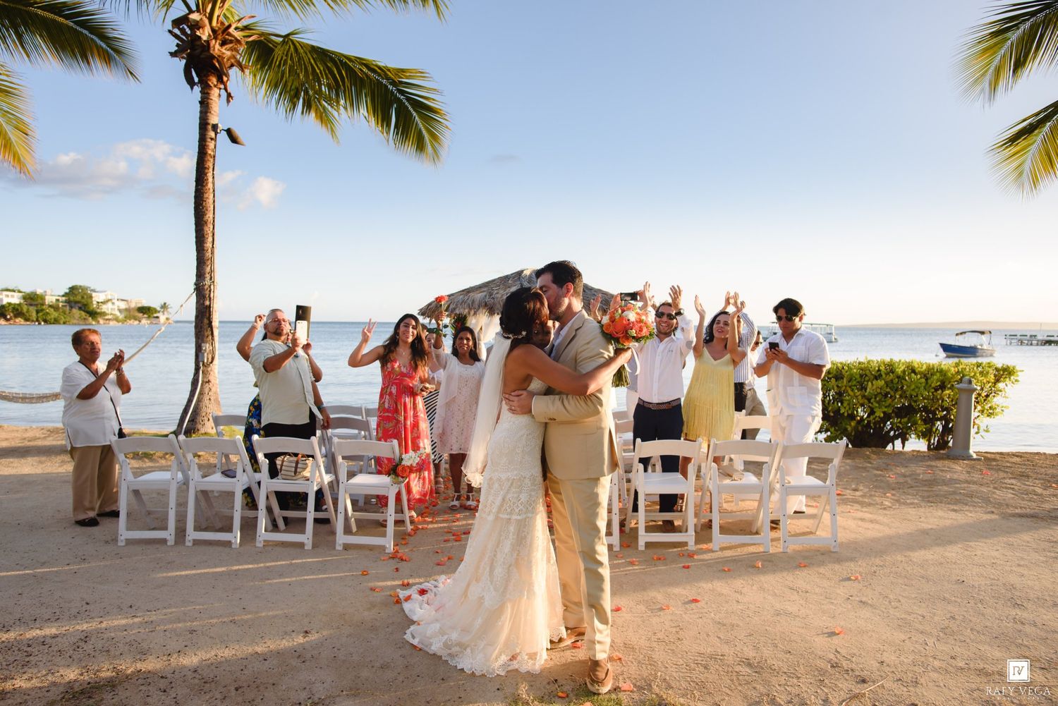 Couple shares first kiss at tropical beach wedding ceremony with guests applauding near palm trees and ocean backdrop.