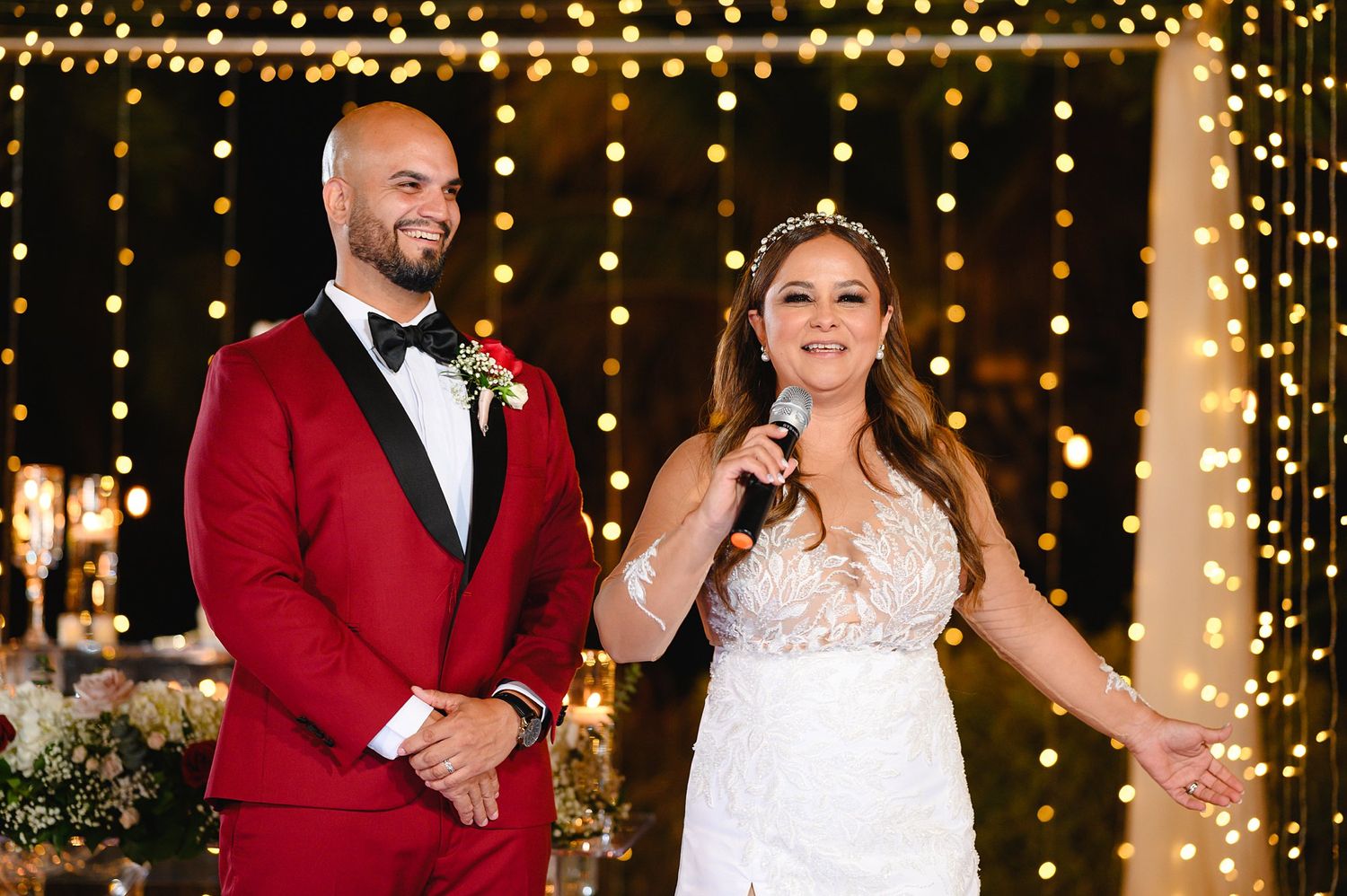 Bride in lace gown and groom in red suit speak at wedding reception with bokeh light backdrop.