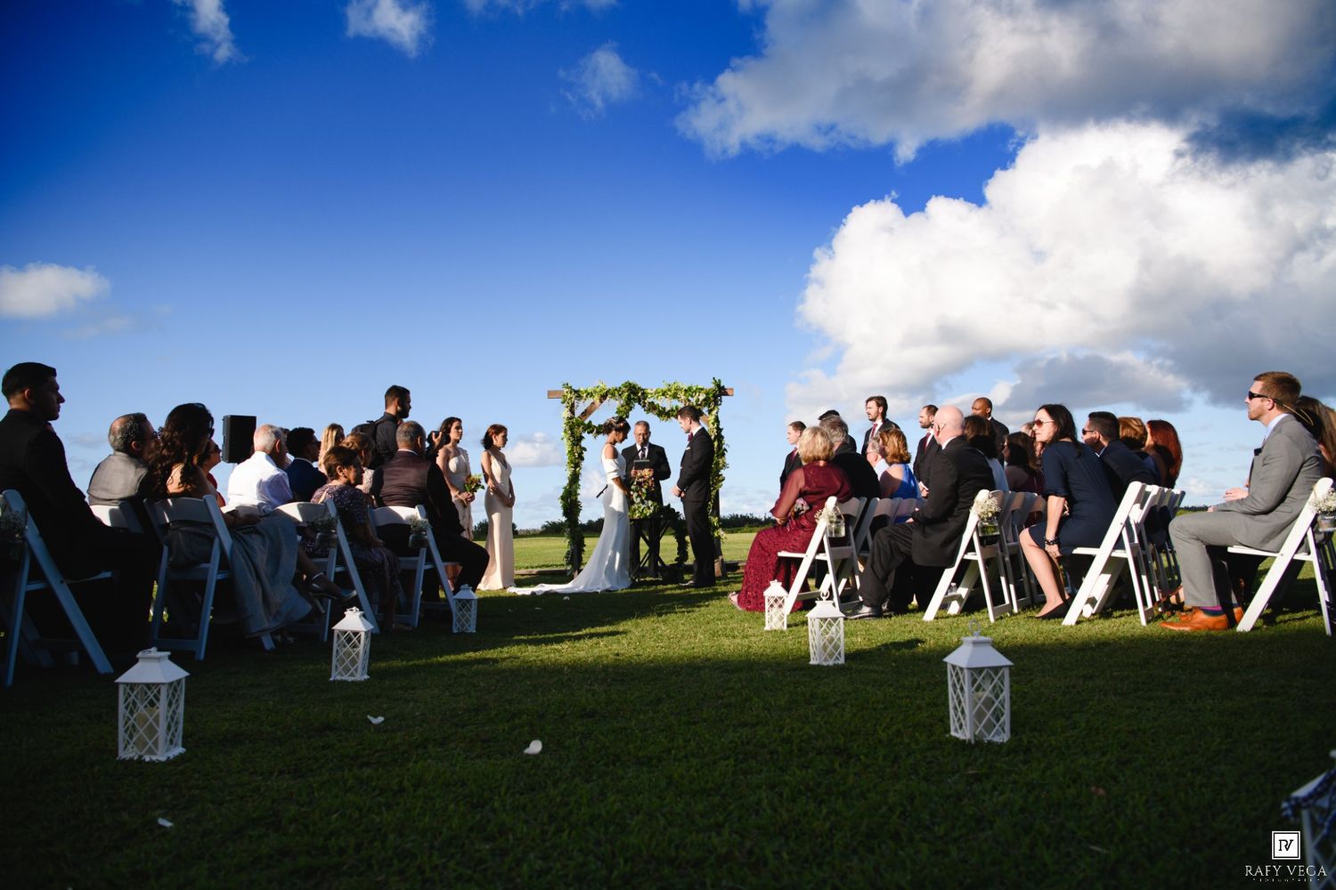 Outdoor wedding ceremony on green lawn with white chairs, floral arch, and blue sky with clouds.