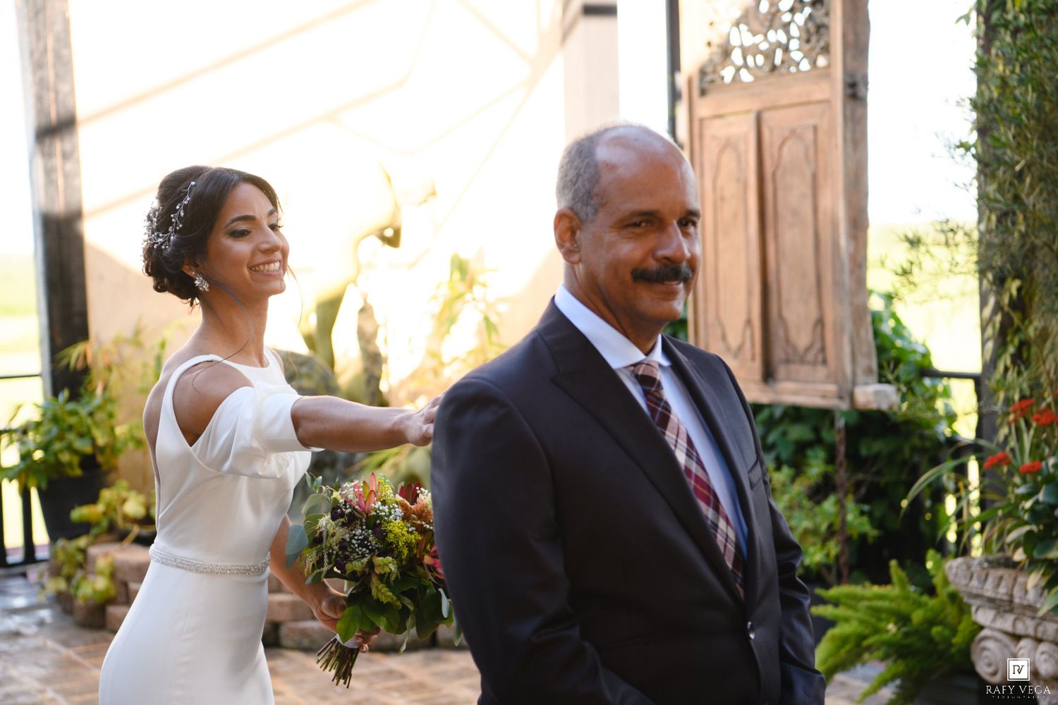 Bride in white gown smiles holding bouquet beside older man in suit at garden wedding venue.