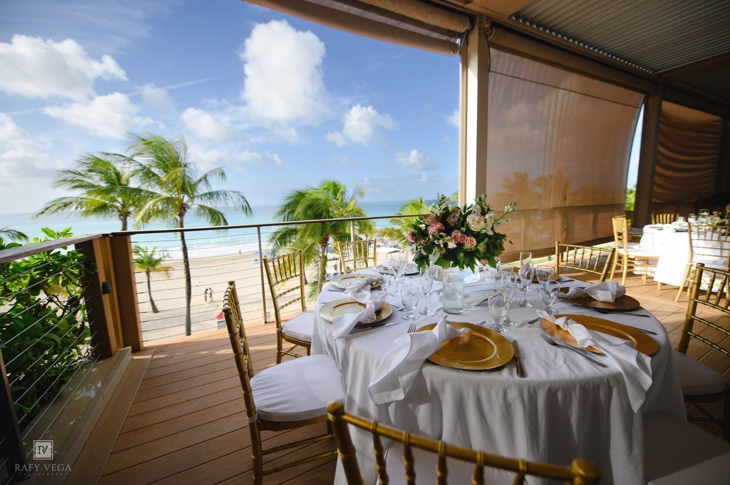 Elegant beachfront wedding reception setup with white tablecloths, gold chargers, and floral centerpieces overlooking palm trees.