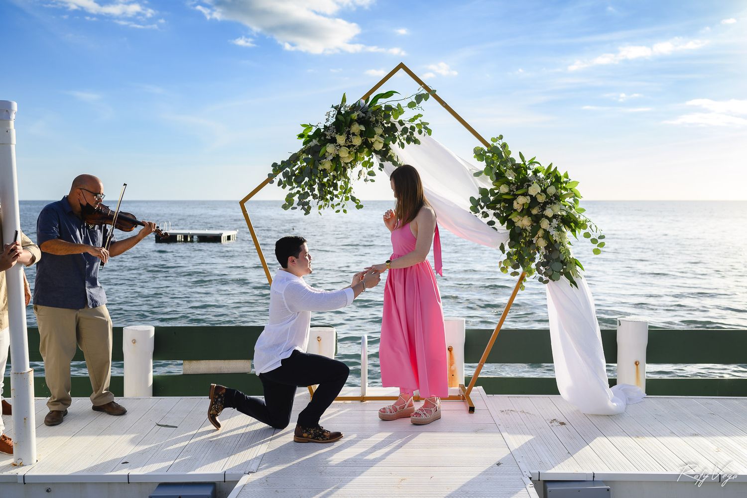 Man proposes to woman in pink dress on waterfront deck with floral arch and violinist at sunset.