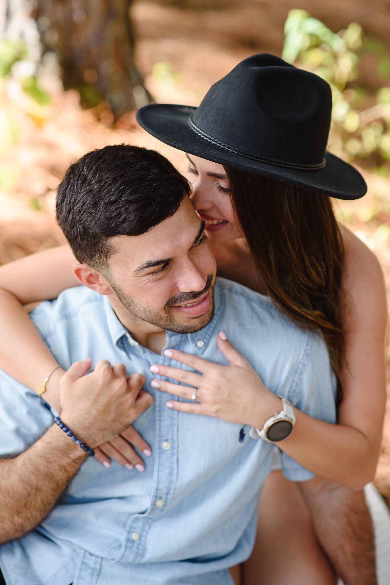 A couple embracing outdoors, woman in black hat kissing man's cheek while he smiles, wearing a blue shirt.