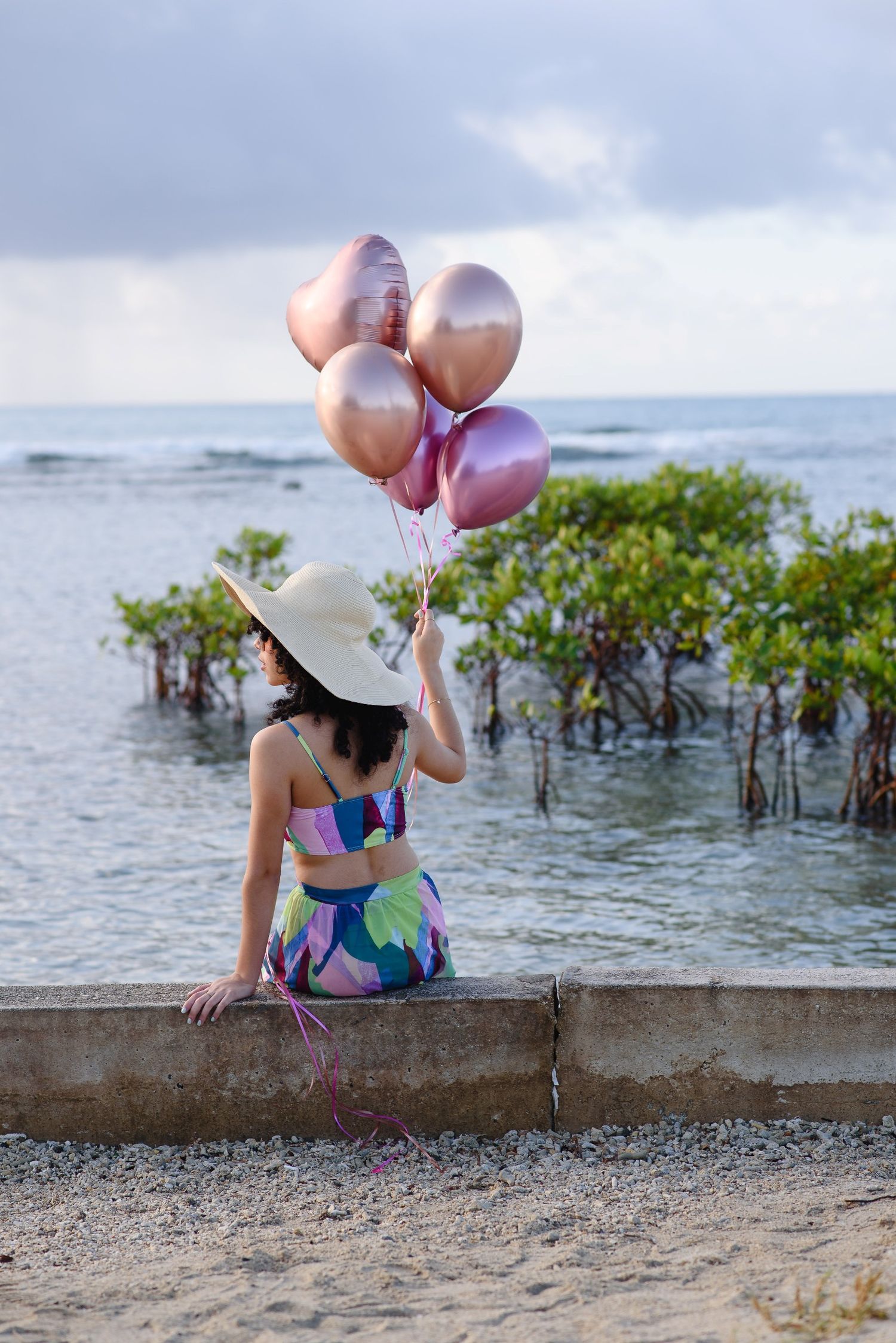 Woman in colorful outfit and white hat sits on beach wall holding pink rose gold balloons near mangroves.