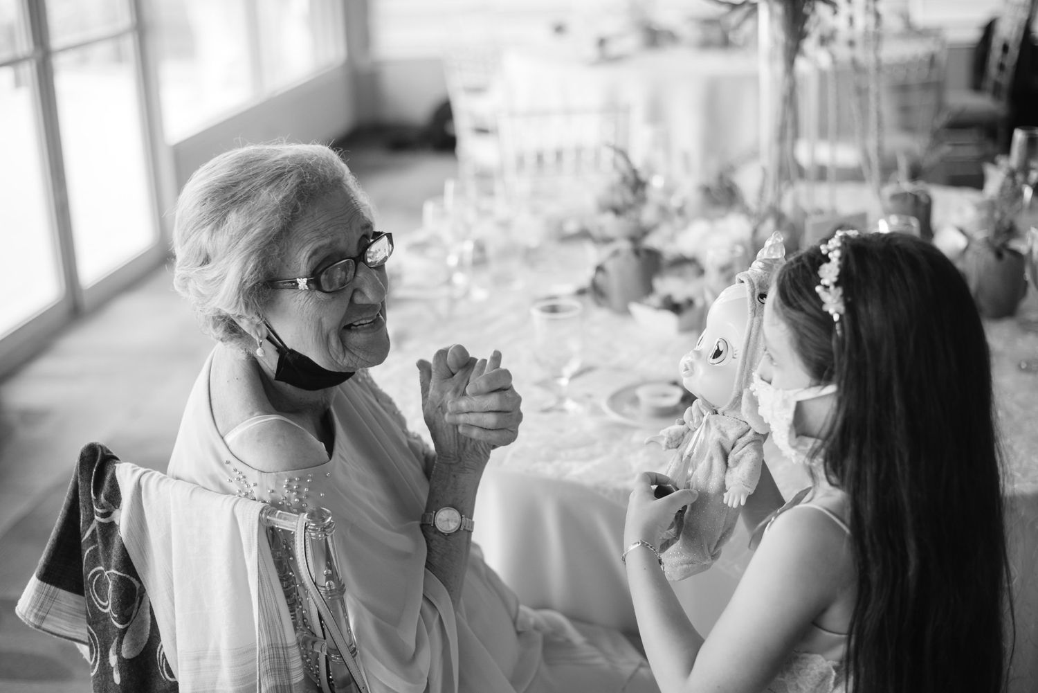Black and white photo of elderly woman and young girl in flower crown sharing a joyful moment at a wedding reception.
