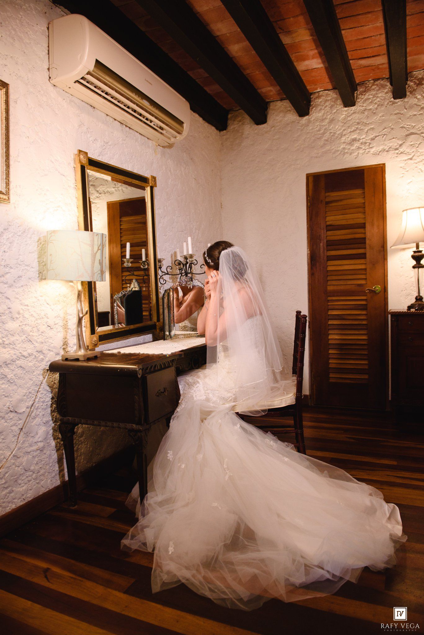 Bride in white mermaid gown with veil sits at vintage vanity mirror in rustic room on wedding day.
