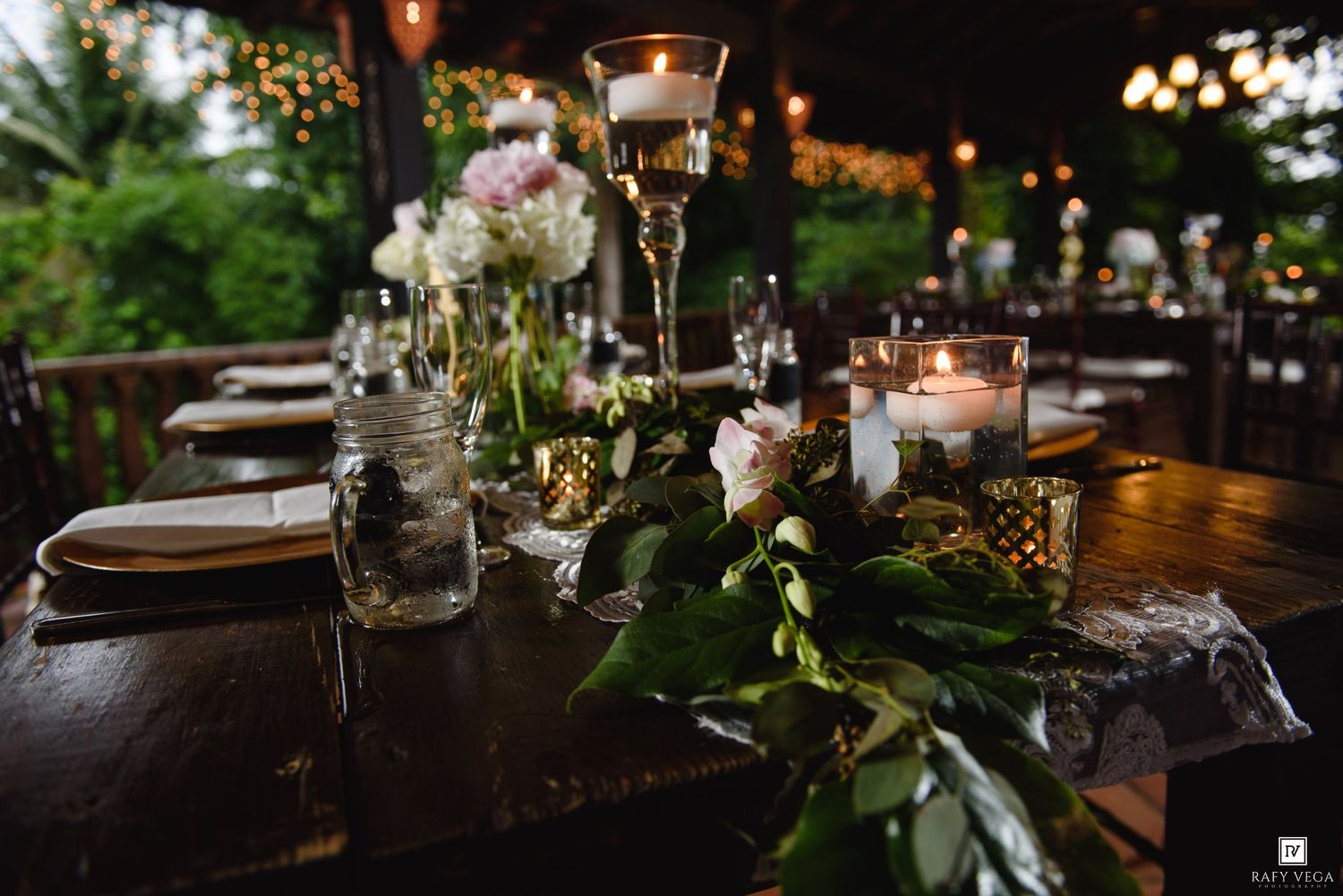 Elegant wedding reception table with candles, floral centerpieces, and greenery garland in an outdoor garden setting.