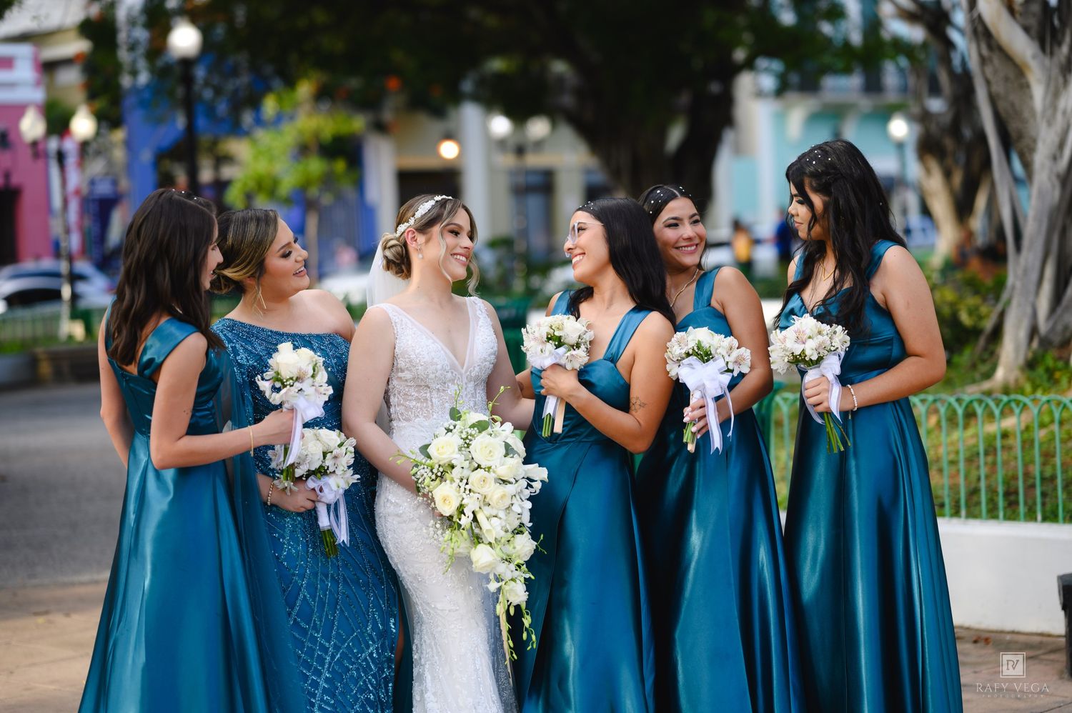 Bride in white gown with bridesmaids in teal satin dresses holding bouquets outdoors.
