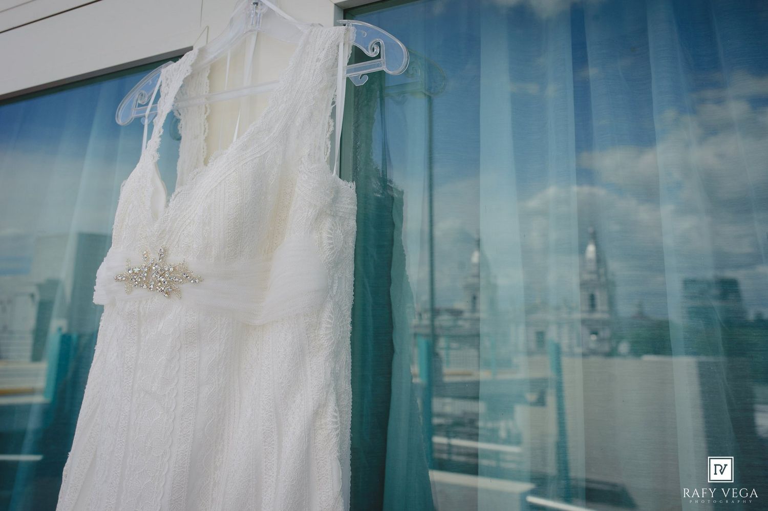 White wedding dress hanging on a glass window with a city skyline reflection, featuring a beaded belt detail.