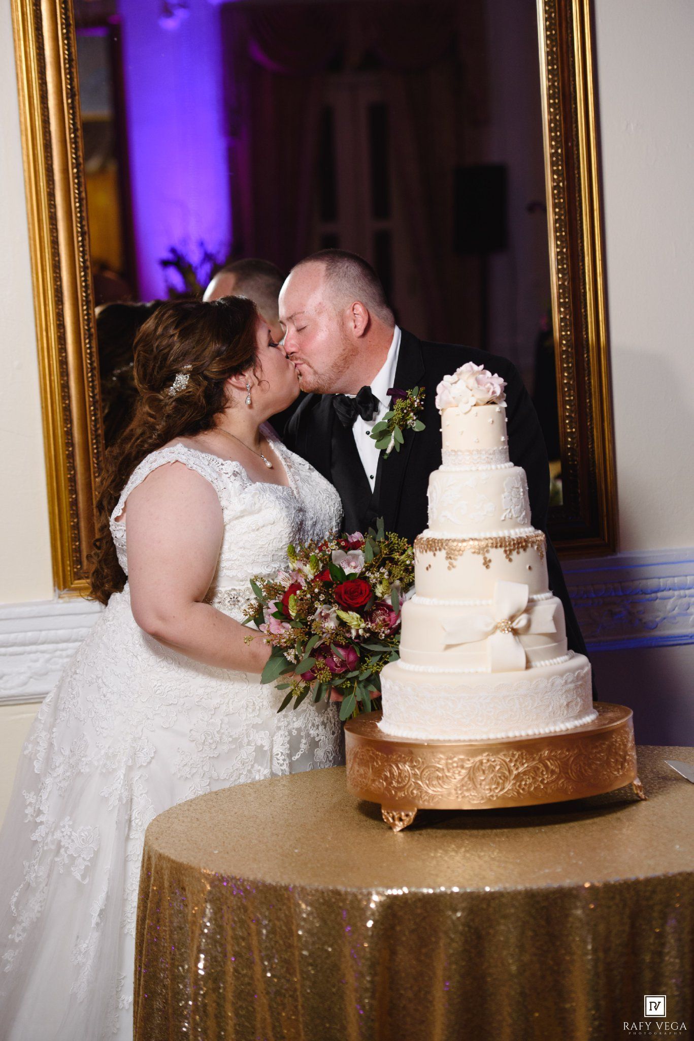 Bride and groom share a kiss beside a elegant white tiered wedding cake on a gold sequin table.