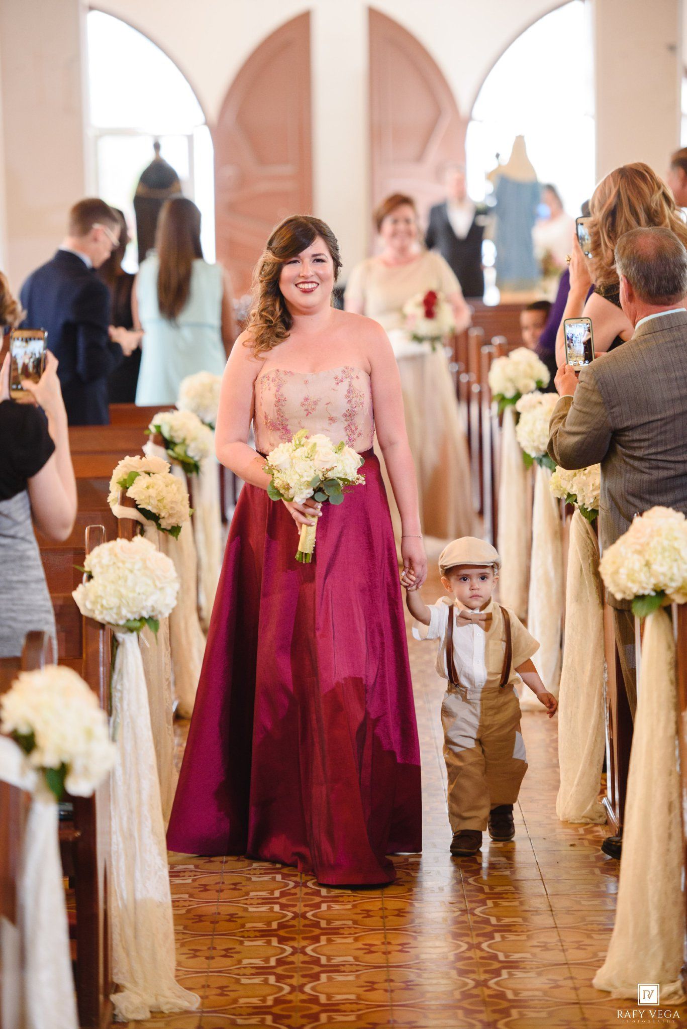 Bridesmaid in burgundy gown walks ring bearer down church aisle holding white bouquet at wedding ceremony.