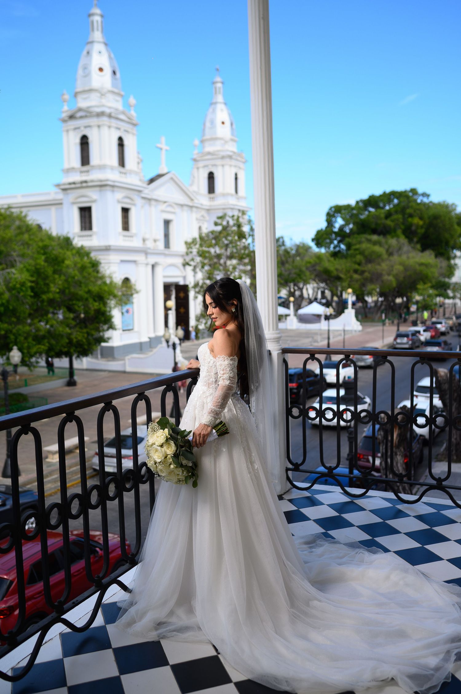 Bride in lace off-shoulder gown with veil poses on balcony with white cathedral in background.