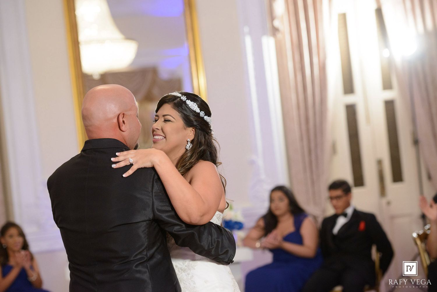 Bride and groom share their first dance at elegant wedding reception with guests in blue attire watching in background.