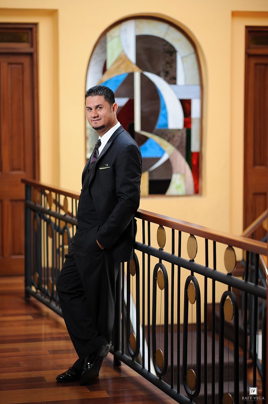 Man in black suit posing on staircase railing with colorful stained glass window backdrop.