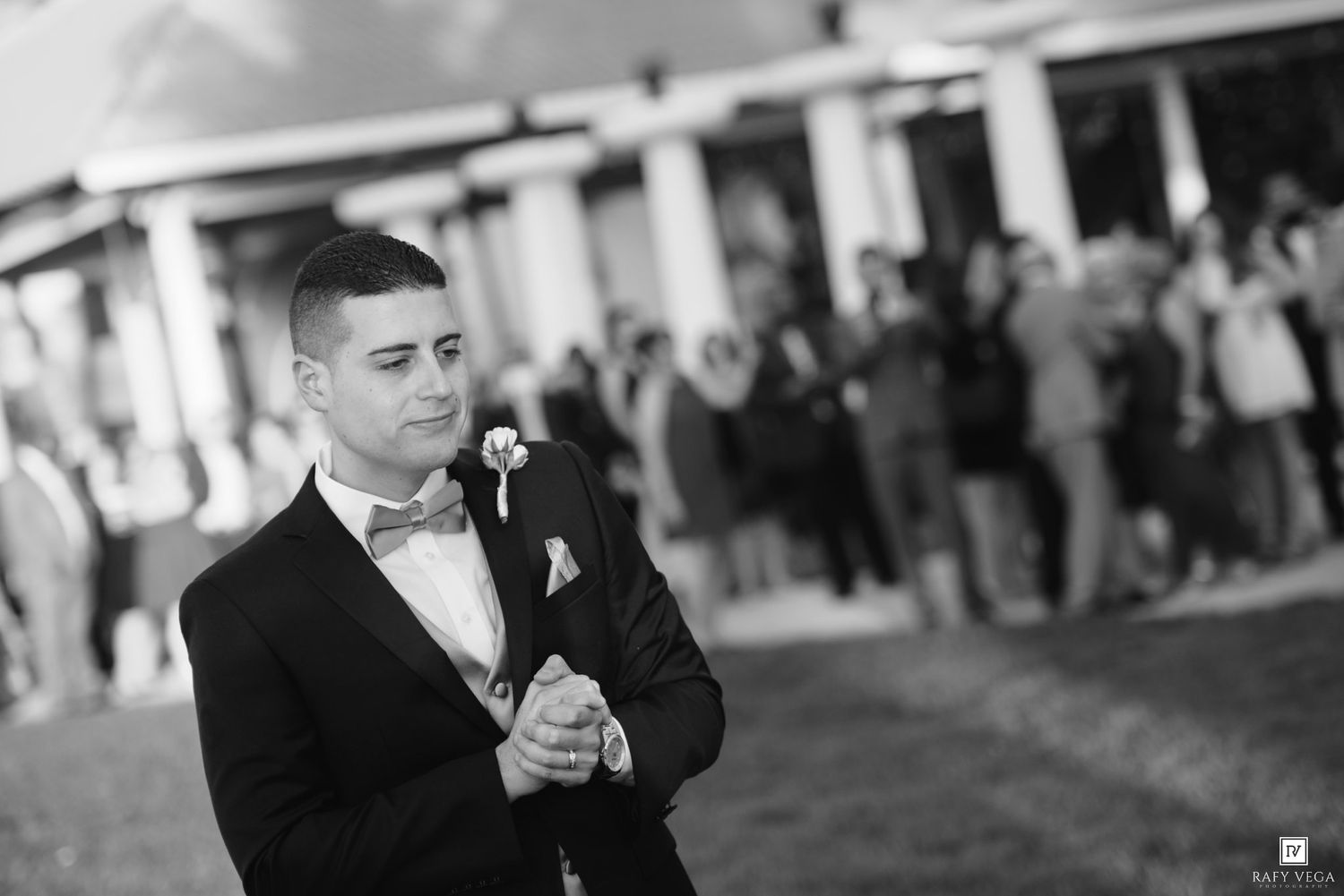Groom in black tuxedo with bow tie stands outdoors at wedding venue with guests in background.